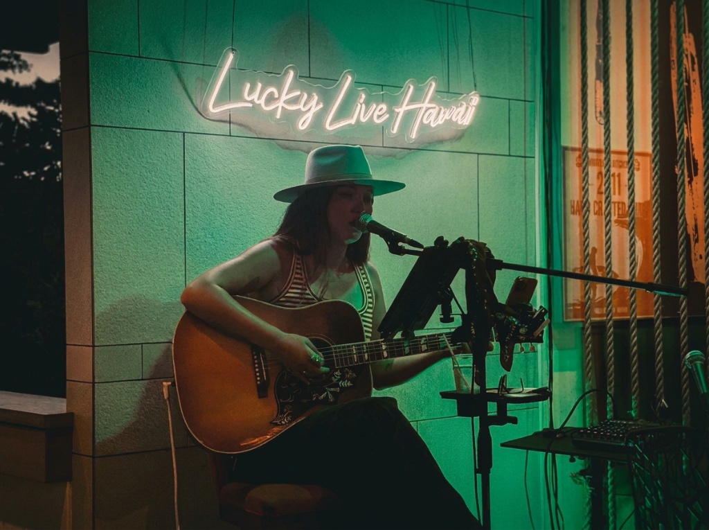 Kris Bradley singing and playing acoustic guitar live at Monkeypod, lit in green neon under a "Lucky Live Hawai'i" sign.