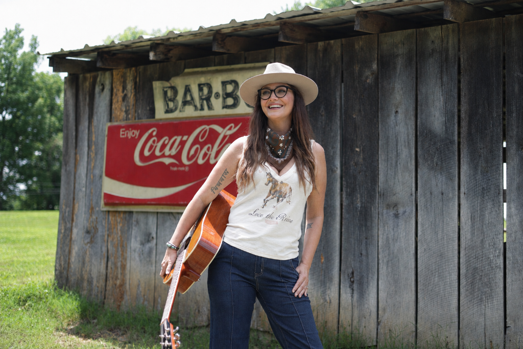 Kris Bradley smiling and playing acoustic guitar in front of a rustic barn.