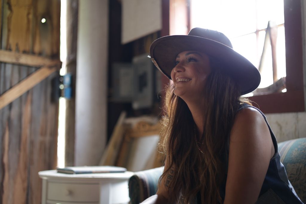 Kris Bradley smiling, lit by warm window light, wearing a wide-brimmed hat.