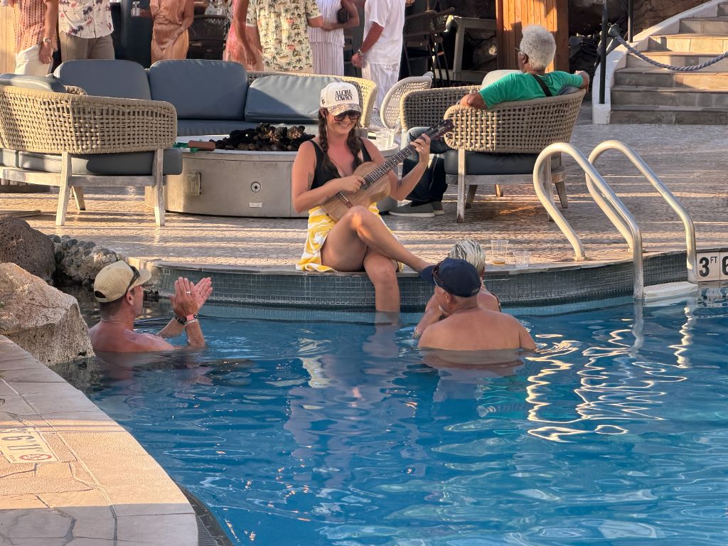 Kris Bradley playing ukulele poolside at a resort while friends look on from the water.